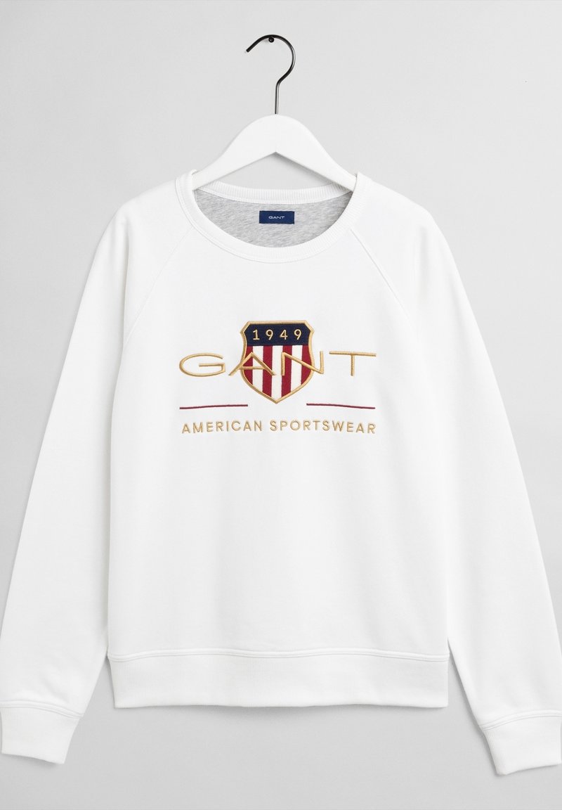Sweat-shirt blanc à manches longues avec col rond, patch logo indiquant "1949 GANT" et texte "American Sportswear" sur le devant, suspendu sur un cintre blanc.