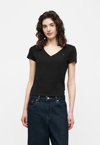 Γυναίκα με καστανά κατσαρά μαλλιά που φορά ένα εφαρμοστό μαύρο T-shirt με V λαιμόκοψη και σκούρο μπλε τζιν, μπροστά σε ένα απλό λευκό φόντο.