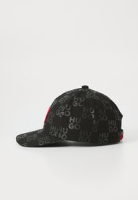 Casquette noire avec un motif « HUGO » répété en finition brillante, présentant un accent rouge solide et une visière structurée. Tissu texturé, arrière ajustable.