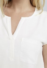 Biała bluzka z krótkim rękawem, z dekoltem w serek, z klapką i małą kieszonką po lewej stronie, wykonana z miękkiego, gładkiego materiału.