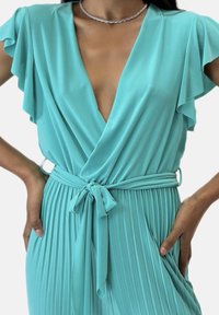 Turkos svepklänning i mjukt tyg, med fjäderärmar, plisserad kjol och knytband i midjan.