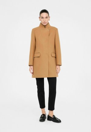 Cappotto in lana marrone con collo alto, due tasche frontali e taglio dritto. Indossato sopra pantaloni neri aderenti e mocassini neri.