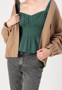 Giacca marrone chiaro con zip e maniche imbottite sopra una maglia verde plissettata. Jeans grigi a vita alta. Focus sulla texture e sul design a strati.
