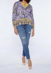 Fialový vzorovaný top se zlatými akcenty, výstřihem do V a průstřihem, v kombinaci s potrhanými modrými džínami a béžovými špičatými lodičkami.