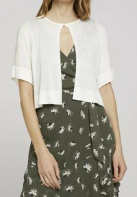 Cardigan bianco corto con maniche corte, caratterizzato da un'apertura a chiave nella parte anteriore e una texture liscia, indossato sopra un vestito floreale verde.