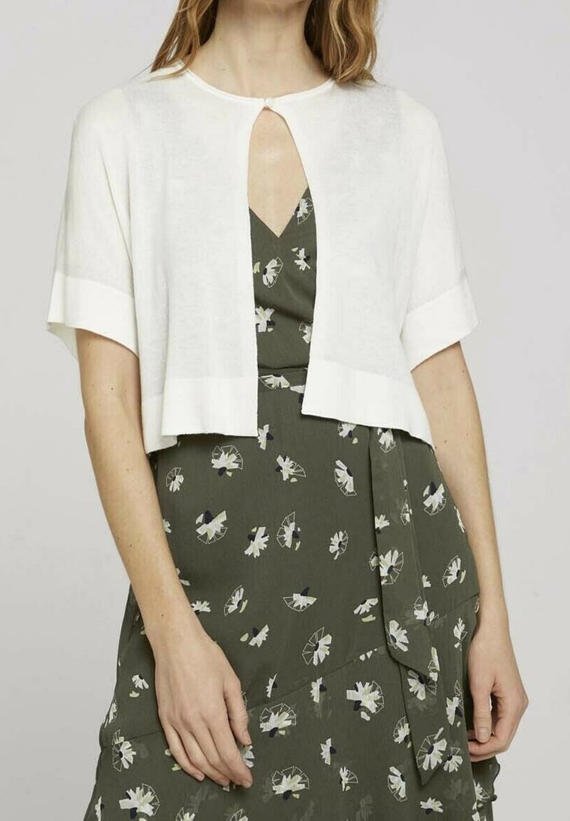 Cardigan bianco corto con maniche corte, caratterizzato da un'apertura a chiave nella parte anteriore e una texture liscia, indossato sopra un vestito floreale verde.