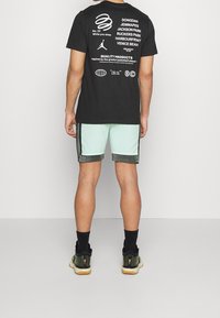 Svart t-shirt med vit grafisk text på ryggen, kombinerad med mintgröna shorts med svart kant och grå texturerade detaljer, samt svarta sneakers.