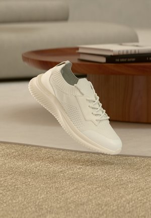 Baskets en tricot blanc avec lacets flottant au-dessus d'un tapis beige tissé, table basse en bois avec des livres empilés sur un sol clair en arrière-plan.