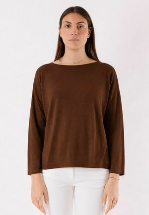 Donna con lunghi capelli scuri che indossa una maglia marrone a maniche lunghe e pantaloni bianchi, in piedi davanti a uno sfondo chiaro e uniforme.