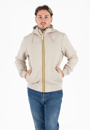 Beige zip-up hoodie met een trekkoord capuchon, voorzien van gele en oranje accenten langs de rits, en twee frontzakken. Draagt met een blauwe spijkerbroek.