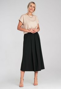Beige Bluse mit kurzen Ärmeln und lockerer Passform, kombiniert mit einem schwarzen, hoch taillierten Plisseerock in Midi-Länge. Klare Absätze runden das Outfit ab.