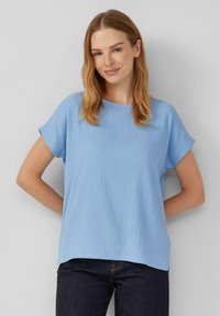 Lichtblauw T-shirt met korte mouwen en een gestructureerd, geribbeld ontwerp; ronde halslijn; relaxte pasvorm; gecombineerd met donkere jeans, handen op de heupen.