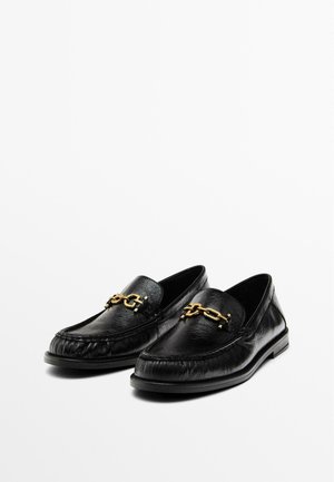 Black leather loafers with a slightly textured finish, featuring a gold chain accent across the front, flat sole, and slip-on design.