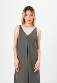 Młoda kobieta z długimi blond włosami, ubrana w bezrękawową ciemnoszaro V-neck sukienkę, stojąca na jednolitym białym tle.