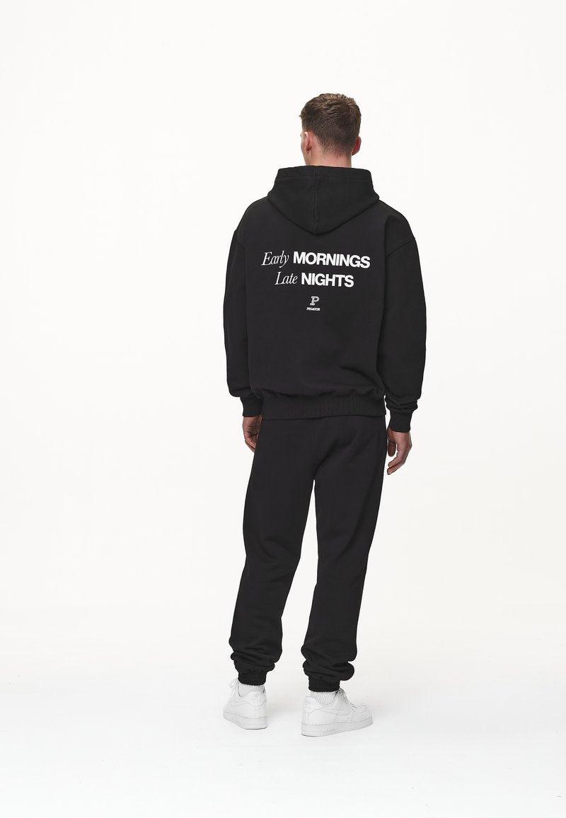 Homme portant un sweat à capuche noir et un pantalon avec le texte blanc "Early MORNINGS Late NIGHTS", debout de dos sur fond blanc.