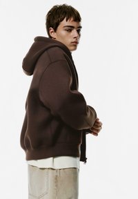 Bruine hoodie met een oversized pasvorm, ribbel manchetten en een rits aan de voorkant. Gelaagd over een crèmekleurige trui en lichtbeige jeans.
