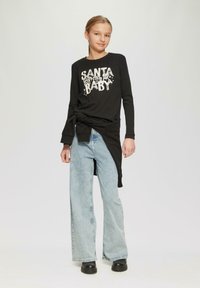 Zwarte lange mouwen shirt met "SANTA BABY" graphic, gecombineerd met lichtblauwe flared jeans en zwarte platformlaarzen. Heeft een gestrikt taille detail.