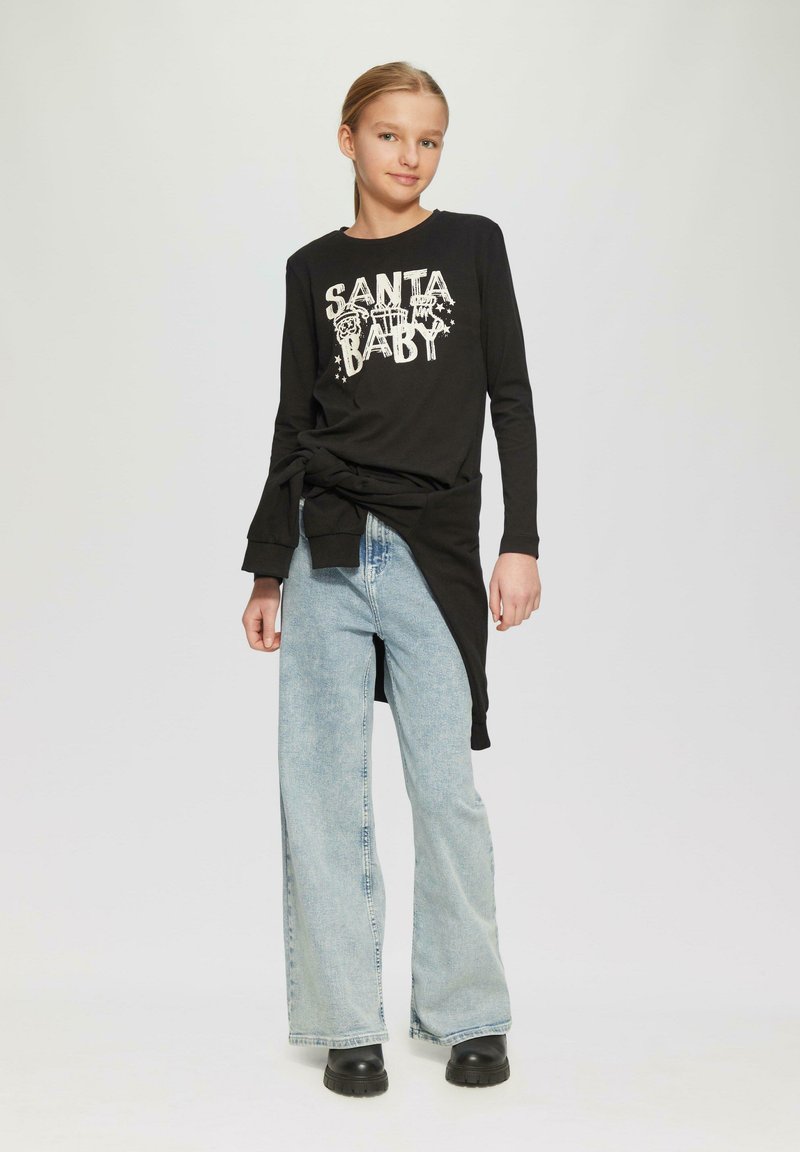 Zwarte lange mouwen shirt met "SANTA BABY" graphic, gecombineerd met lichtblauwe flared jeans en zwarte platformlaarzen. Heeft een gestrikt taille detail.