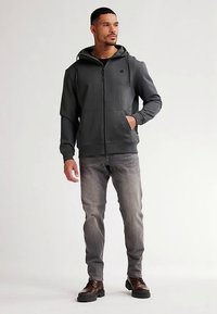 Felpa con cappuccio grigio scuro con zip e coulisse, dotata di tasche laterali e tessuto strutturato, abbinata a jeans grigio chiaro e stivali.