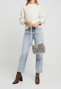 Kobieta w kremowym swetrze w warkocze, jasnoniebieskich dżinsach, brązowych butach w wężową skórę i trzymająca czarną torbę z dużym łańcuchem.