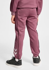 Lyserøde træningsbukser med sorte sidelæg, elastiske manchetter og en chevronlogo-detalje i sølv. Båret med sorte sneakers og hvide sokker.