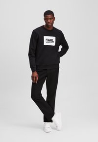 Hanorac negru cu panou frontal alb cu brand, asociat cu pantaloni negri și adidași albi. Design simplu, material din bumbac, guler rotund.