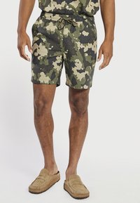 Male wearing green and beige camouflage shorts with drawstring waist and tan slip-on loafers against plain background.