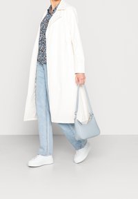 Un leggero cappotto bianco sopra una camicia floreale, abbinato a jeans azzurri e sneakers bianche. Una piccola borsa a spalla azzurra completa il look.