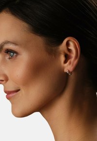 Profilo laterale di una donna che mostra una pelle liscia, capelli scuri raccolti e un piccolo orecchino a cerchio dorato con uno smeraldo trasparente.