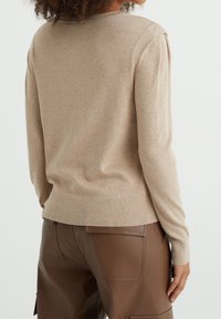 Béžový pletený svetr s ribbovanými manžetami a lemem, s jednoduchým designem a měkkou texturou, nošený s hnědými koženými kalhotami.