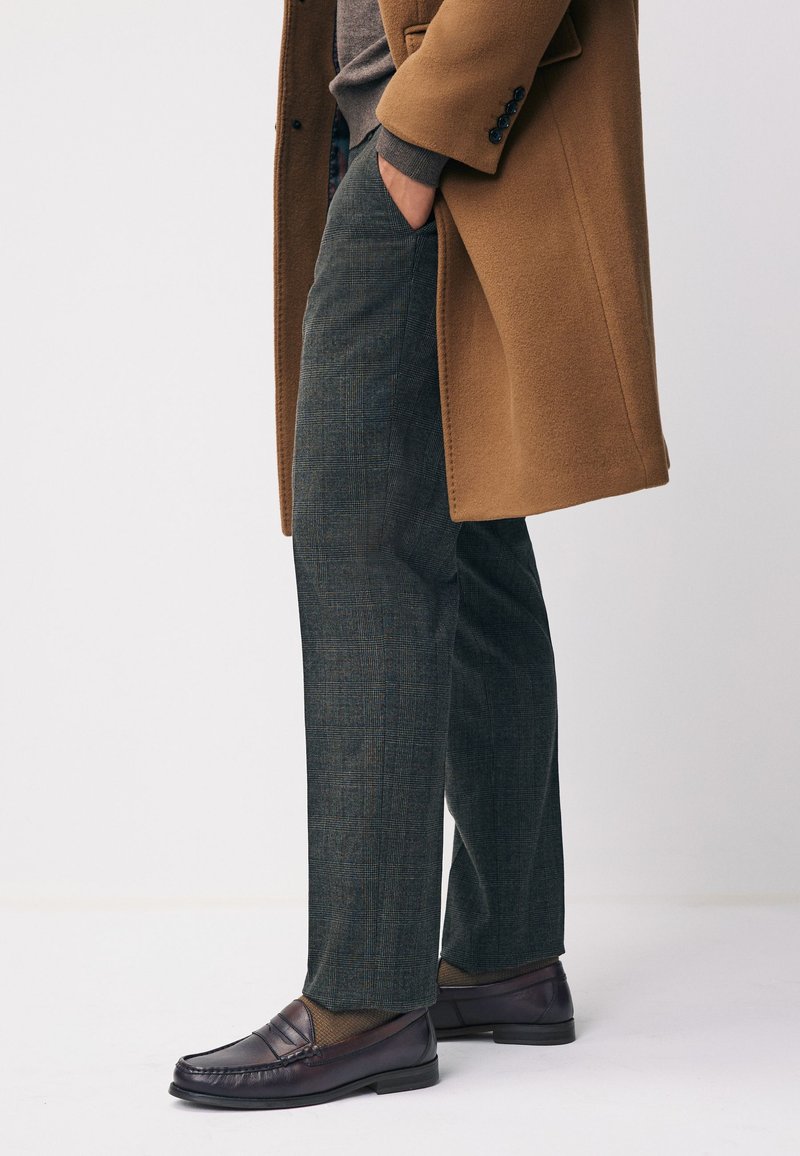 Homme portant un manteau marron, un pantalon à carreaux gris et des mocassins en cuir marron foncé, les mains dans les poches, sur un fond uni.