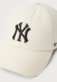 Cream-colored baseball cap with a structured design, featuring a black embroidered "NY" logo on the front and stitched detailing on the brim.