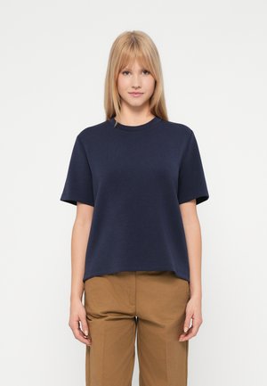 Blonde vrouw staat, gekleed in een marineblauwe shirt met korte mouwen en lichtbruine broek, tegen een plain witte achtergrond.