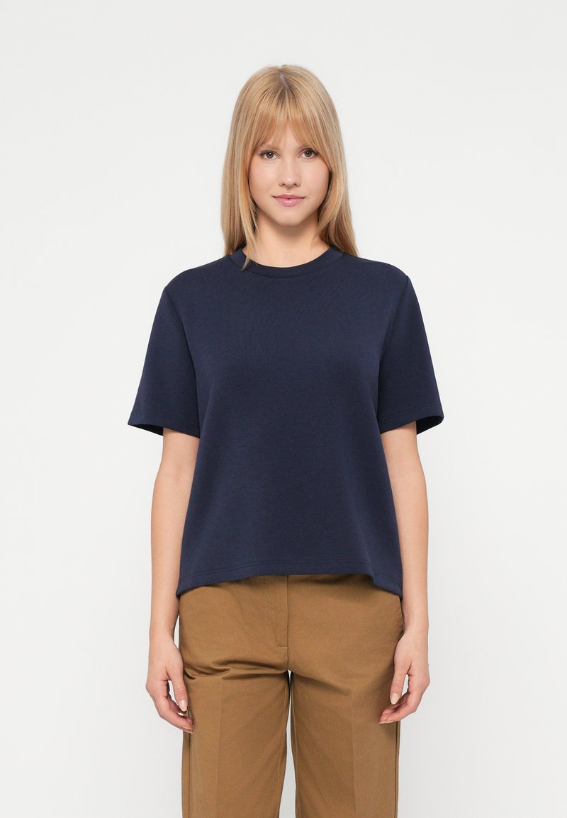 Ξανθιά γυναίκα που στέκεται, φορώντας ένα ναυτικό μπλε κοντομάνικο πουκάμισο και ανοιχτό καφέ παντελόνι, μπροστά από έναν απλό λευκό τοίχο.