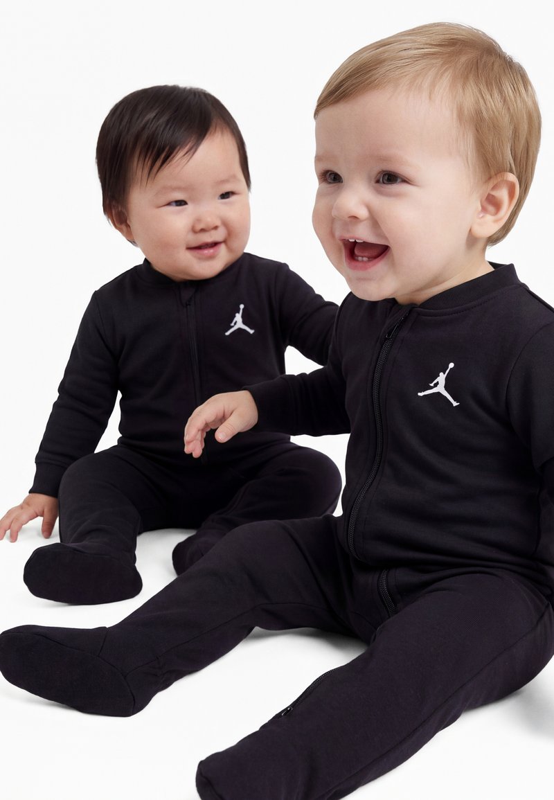 Deux bébés assis et souriants, vêtus de tenues noires assorties zippées avec un logo Jumpman blanc sur la poitrine.