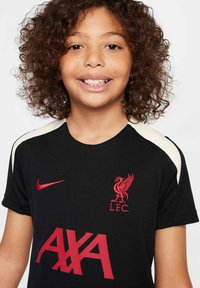 Crna Liverpool FC majica s crvenim logotipom i natpisom "AXA", s krem detaljima na ramenima. Izrađena od teksturirane tkanine.