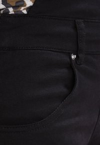 Gros plan sur la poche d'un jean en denim noir avec des coutures visibles, un rivet argenté et un aperçu d'un tissu à imprimé léopard au-dessus de la ceinture.