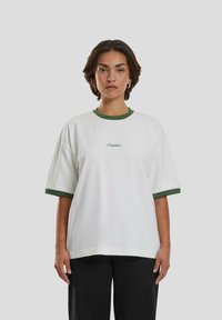 Camiseta blanca de algodón con cuello y dobladillos de las mangas de canalé verde, que presenta la palabra "Prohibido" en un estilo cursivo verde en el pecho.
