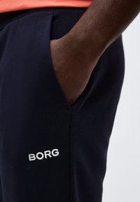 Χέρι στην τσέπη σκούρων μπλε παντελονιών με λευκό λογότυπο «BORG», φοράει πορτοκαλί πουκάμισο, σε απλό γκρι φόντο.