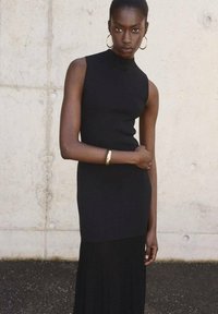 Abito nero senza maniche con collo alto, corpino aderente e gonna plissettata fluente. La modella indossa orecchini a cerchio dorati e un bracciale abbinato.
