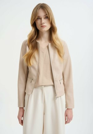 Beige koženka s předním zipem, bočními kapsami a kulatým výstřihem. Nositá přes světle béžový top a krémové kalhoty.