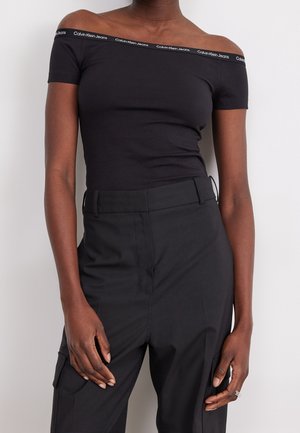 Personne mince portant un haut noir épaules dénudées avec la marque Calvin Klein Jeans et un pantalon noir taille haute sur un fond uni.