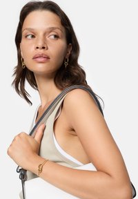 Orecchini a cerchio dorati, un top a coste beige e una borsa bianca con una tracolla nera. Pelle liscia, trucco naturale e capelli ondulati di lunghezza media.