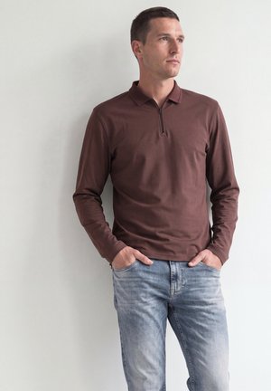 Mann trägt braunes langärmeliges Polohemd mit Reißverschluss und hellblaue Jeans, steht mit den Händen in den Taschen vor einer einfarbigen Wand.