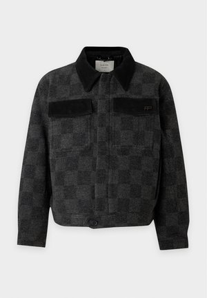 Homme portant une veste à carreaux foncée avec des poches, un col roulé noir, un jean bleu ample et des chaussures noires sur un fond blanc uni.