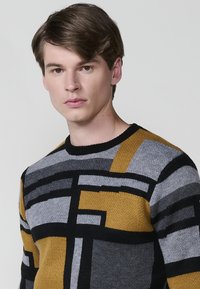 Suéter de punto con patrones geométricos en negro, gris y amarillo mostaza. Diseño de cuello redondo con ribete acanalado y una textura suave.