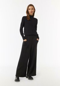 Schwarzer Rollkragenpullover mit langen Ärmeln, kombiniert mit weiten schwarzen Hosen mit seitlichen Taschen und einem Satinstreifen. Getragen mit schwarzen Ankle Boots.