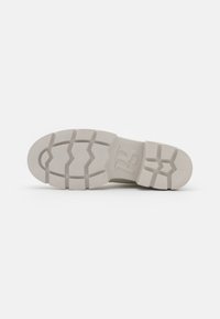 Suela de una zapatilla, con un diseño grueso y texturizado en color crema claro, con surcos contrastantes y un logo prominente en la base.