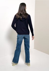 Marineblaue Kapuzenjacke mit Kordelzug, kombiniert mit lockeren, hellblauen Jeans. Weiße Sneakers mit grünen Akzenten. Rückansicht.