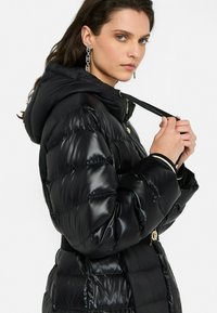 Žena upravuje popruh lesklého čierneho puffa s kapucňou, má na sebe dlhé náušnice z reťazí, na jednoduchom pozadí.